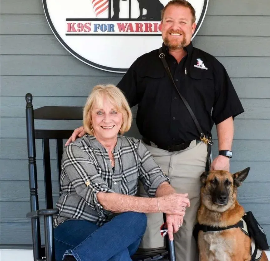 A veteran and a smiling woman pose with a German Shepherd service dog beneath the K9s For Warriors sign